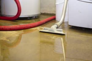 A flooded basement floor with standing water, a wet vacuum hose, and cleaning equipment used for water removal.